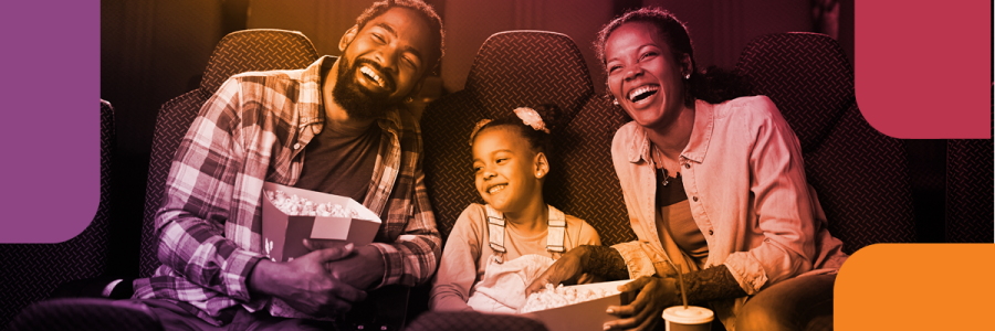 A man, child, and woman laugh as they eat popcorn in a movie theatre.