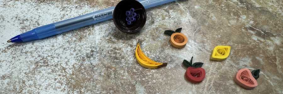 Tiny brown bowl with a banana, orange, apple, lemon, peach, and grapes. Pen shown for scale (tiny!).