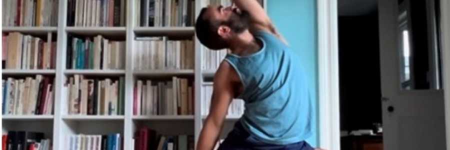 Bearded man in a yoga pose on a mat in a room with bookshelves in the background